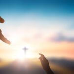 Hand extending from above to a hand reaching up from below; background of sky.